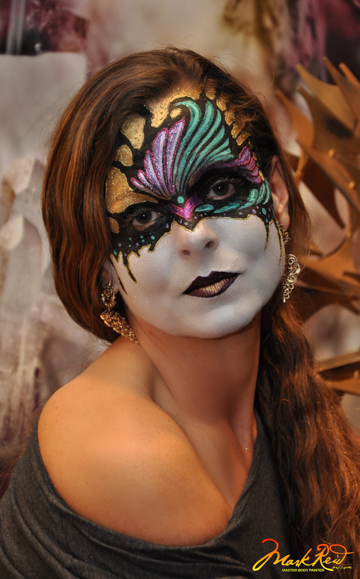 Brunette woman in full face paint that features a white lower half of the face with black lips and a colorful mask on the top half